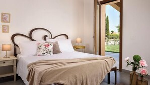 A neatly made bed with a beige blanket, two bedside lamps, and a floral pillow. There is a small table with a lamp beside the bed, and a view of a garden through an open door. - Borgo Verde (Rosignano Marittimo)