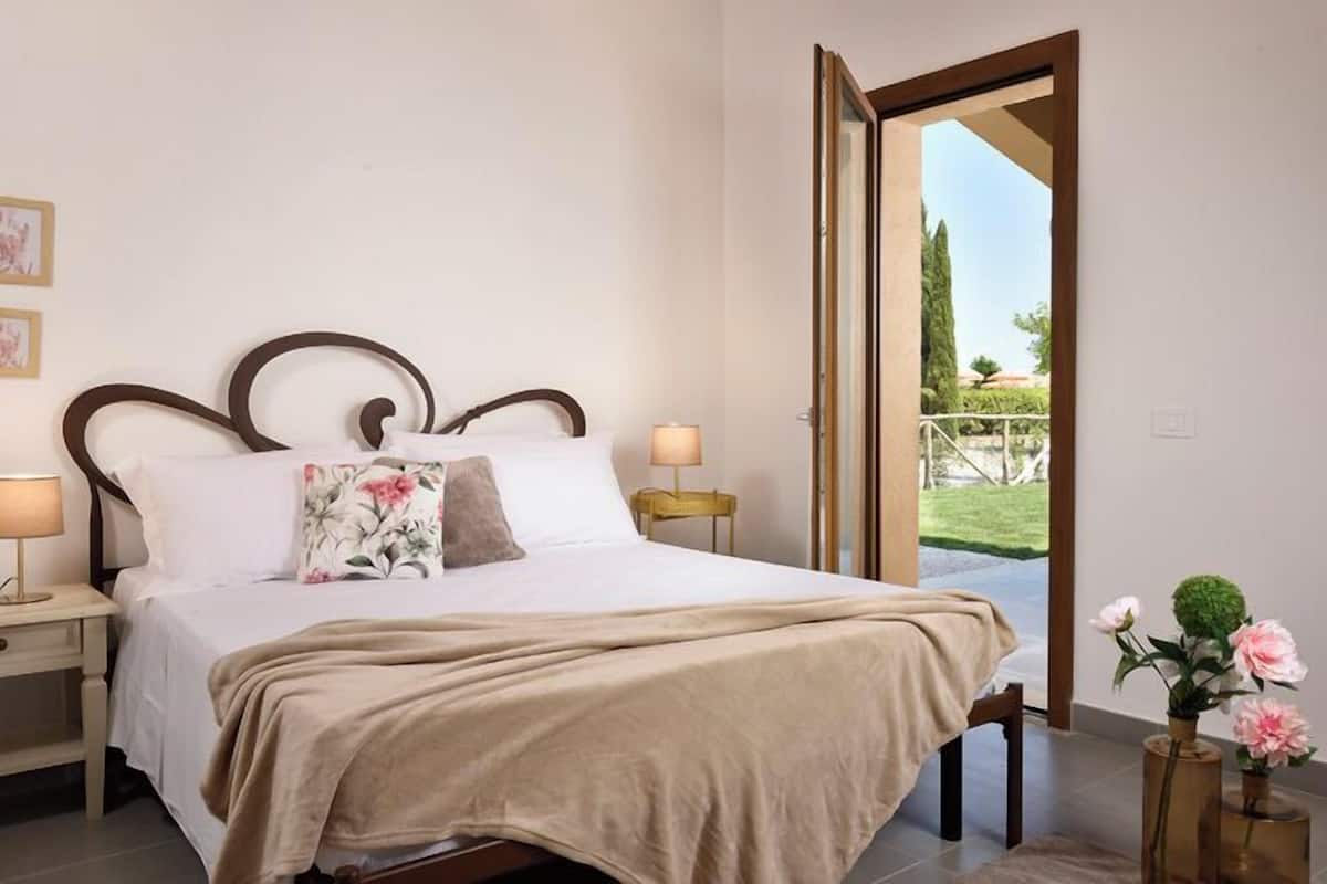 A neatly made bed with a beige blanket, two bedside lamps, and a floral pillow. There is a small table with a lamp beside the bed, and a view of a garden through an open door.