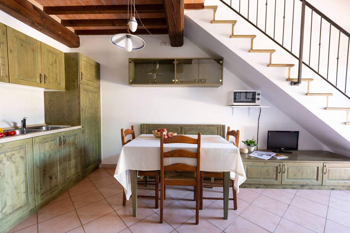 A kitchen with wooden cabinets, a dining table with chairs, a microwave, and a television on a shelf.