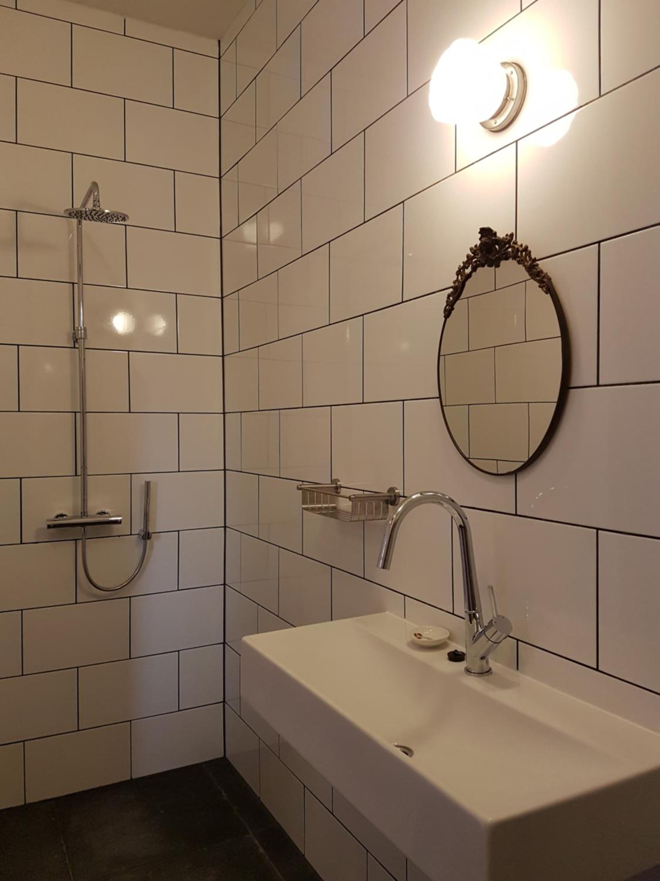 A modern bathroom with white tiles, a rectangular sink, a shower with a handheld showerhead, and a wall-mounted mirror.