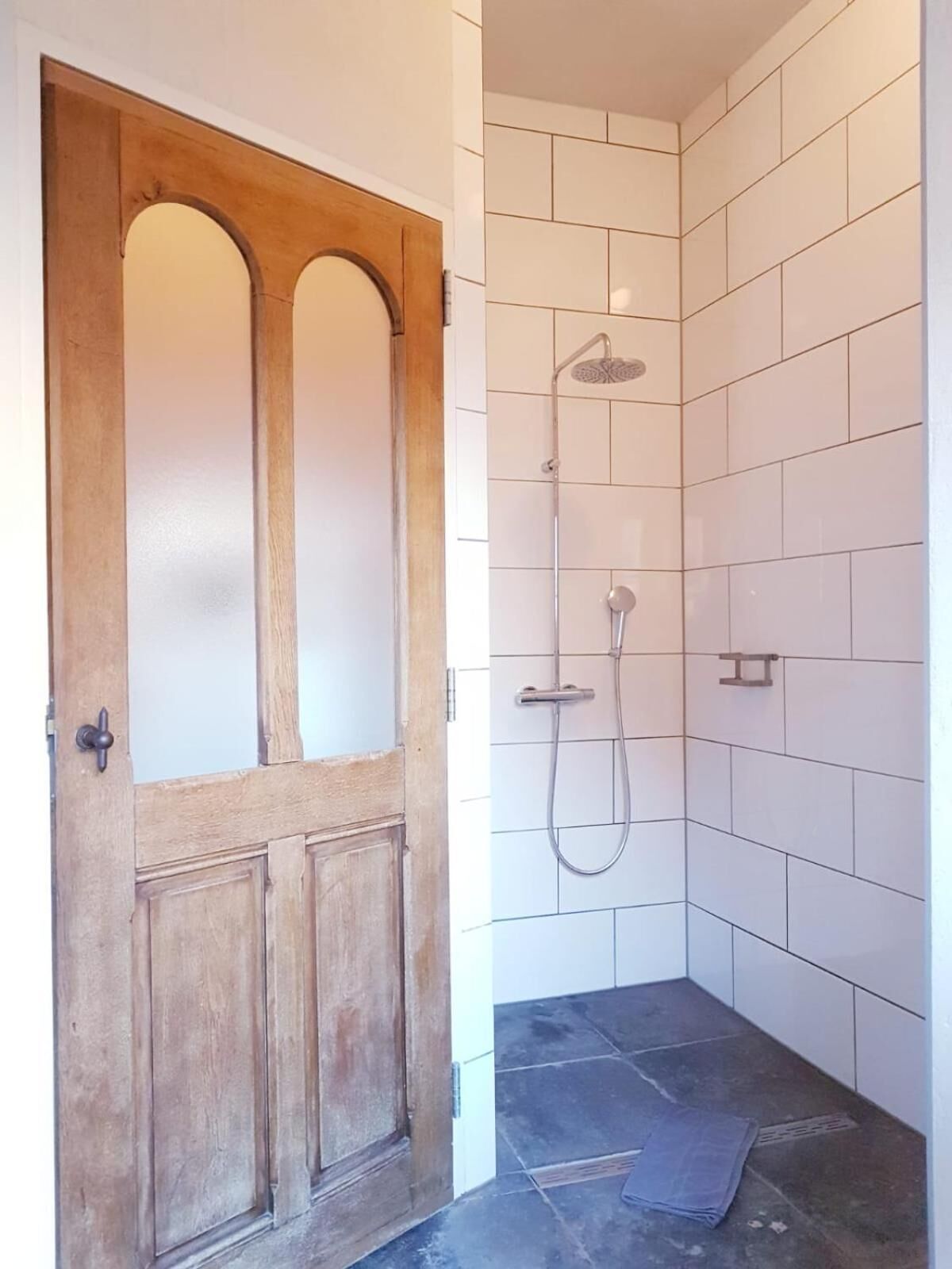 A bathroom with a wooden door, white tiled walls, a shower with a handheld showerhead, and a dark tiled floor.