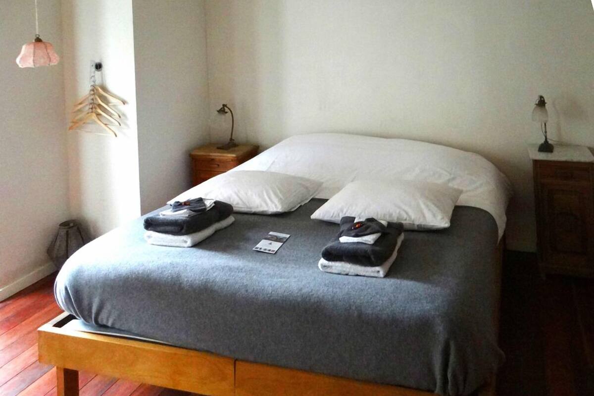 Een net opgemaakt bed met een grijze deken, twee witte kussens en opgevouwen handdoeken. Er staan houten nachtkastjes met lampen, een houten bankje aan het voeteneind van het bed, en een wandrek met daaraan kleding.