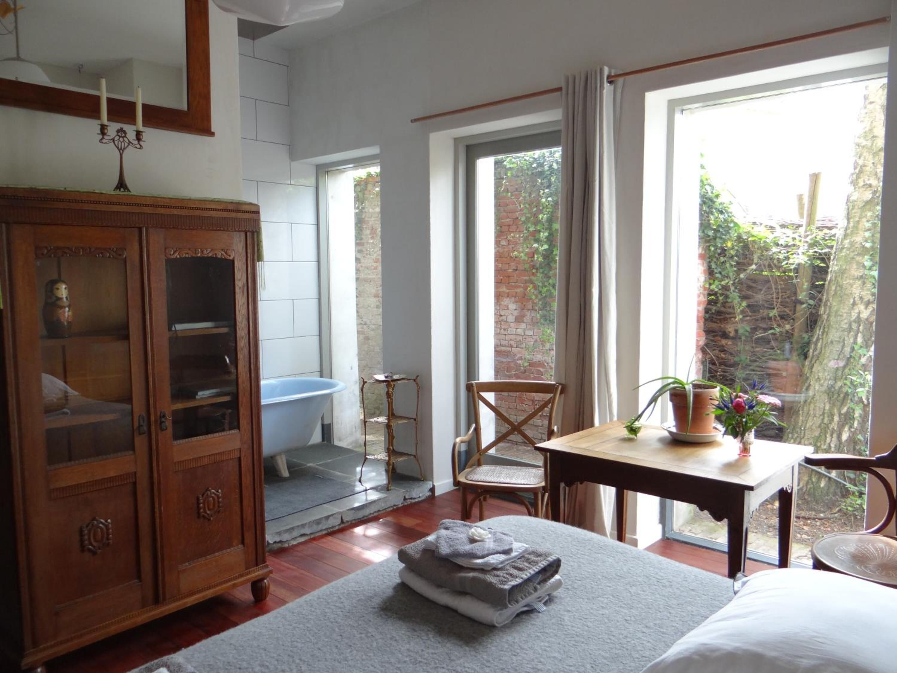 Une chambre avec une grande fenêtre, une armoire en bois, une baignoire et une table sur laquelle est posée une plante.