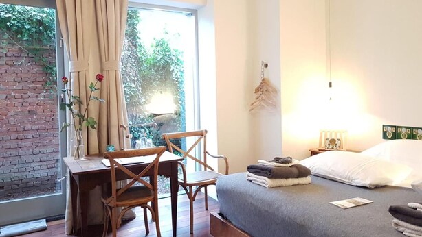 Een slaapkamer met een bed, een bureau en stoelen. Er is een raam met gordijnen, uitzicht op groen buiten, en een wandlamp.