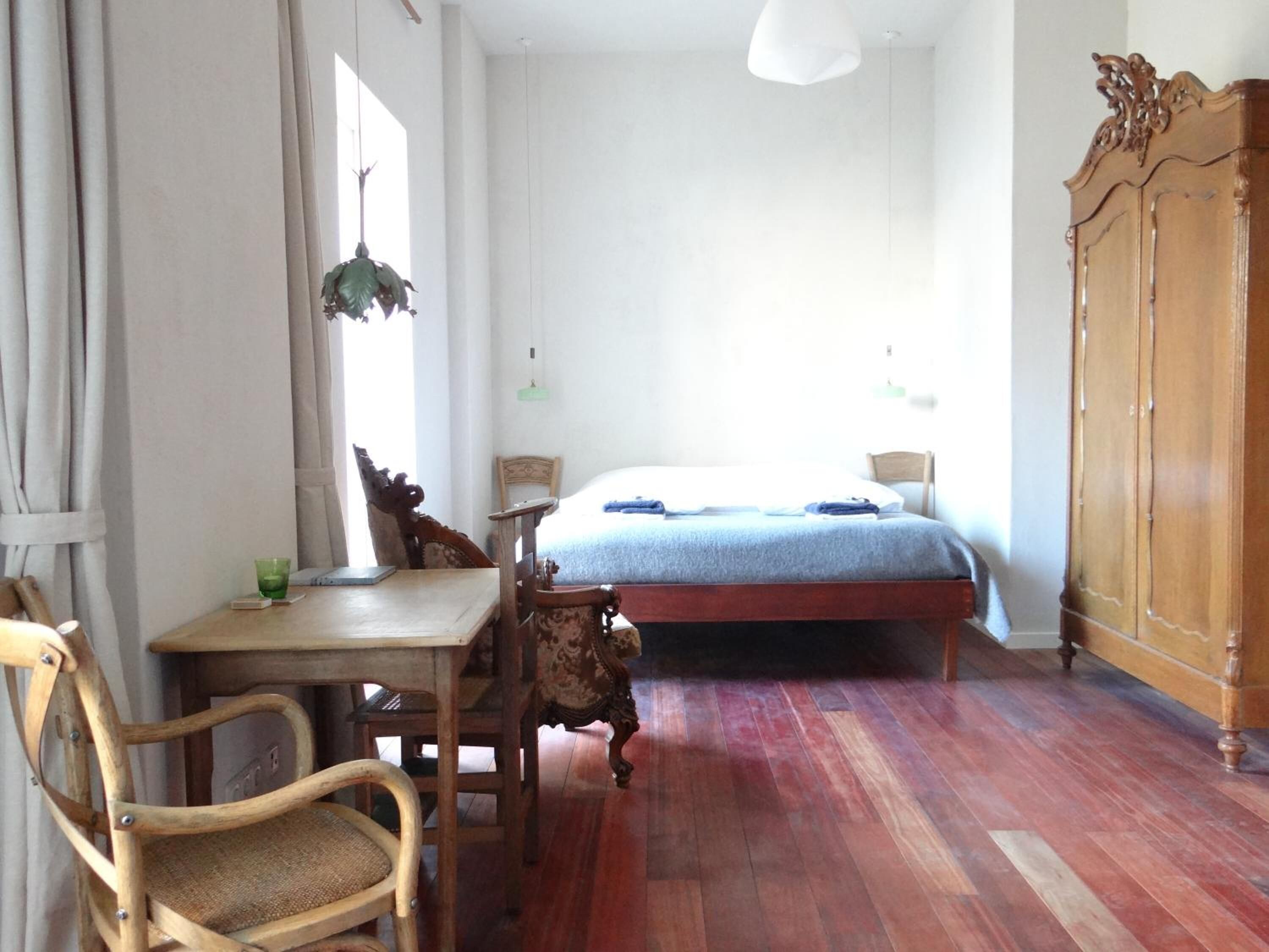 A room with a bed, a wooden wardrobe, a desk with a chair, and a hanging plant.