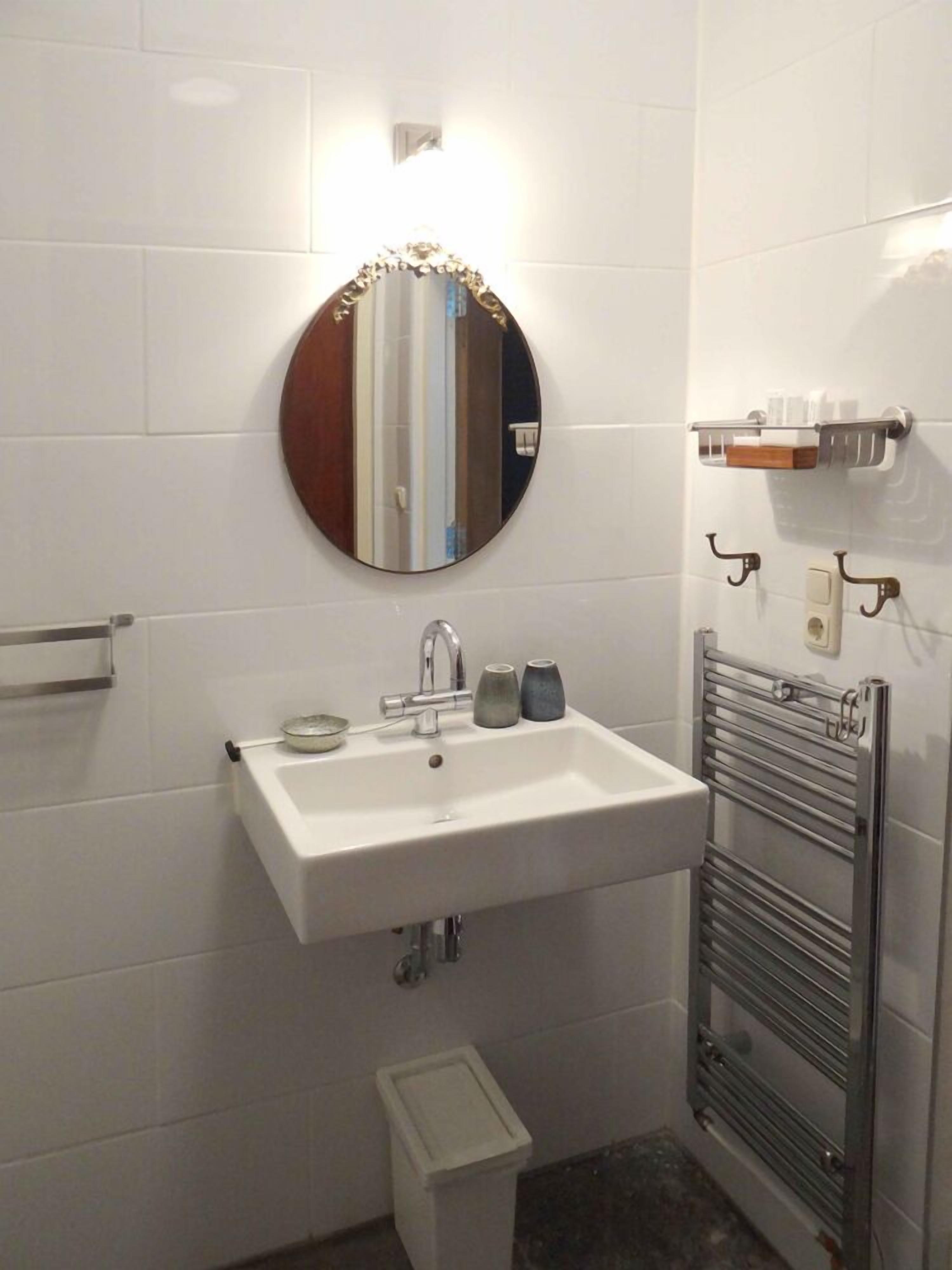 A bathroom with a white sink, a round mirror, a towel rack, and a small trash bin.