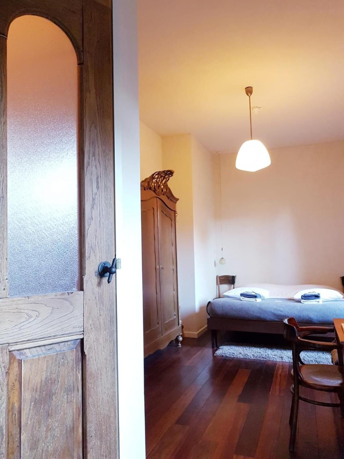 A room with a bed, a wooden wardrobe, a dining table with chairs, and a hanging light fixture.