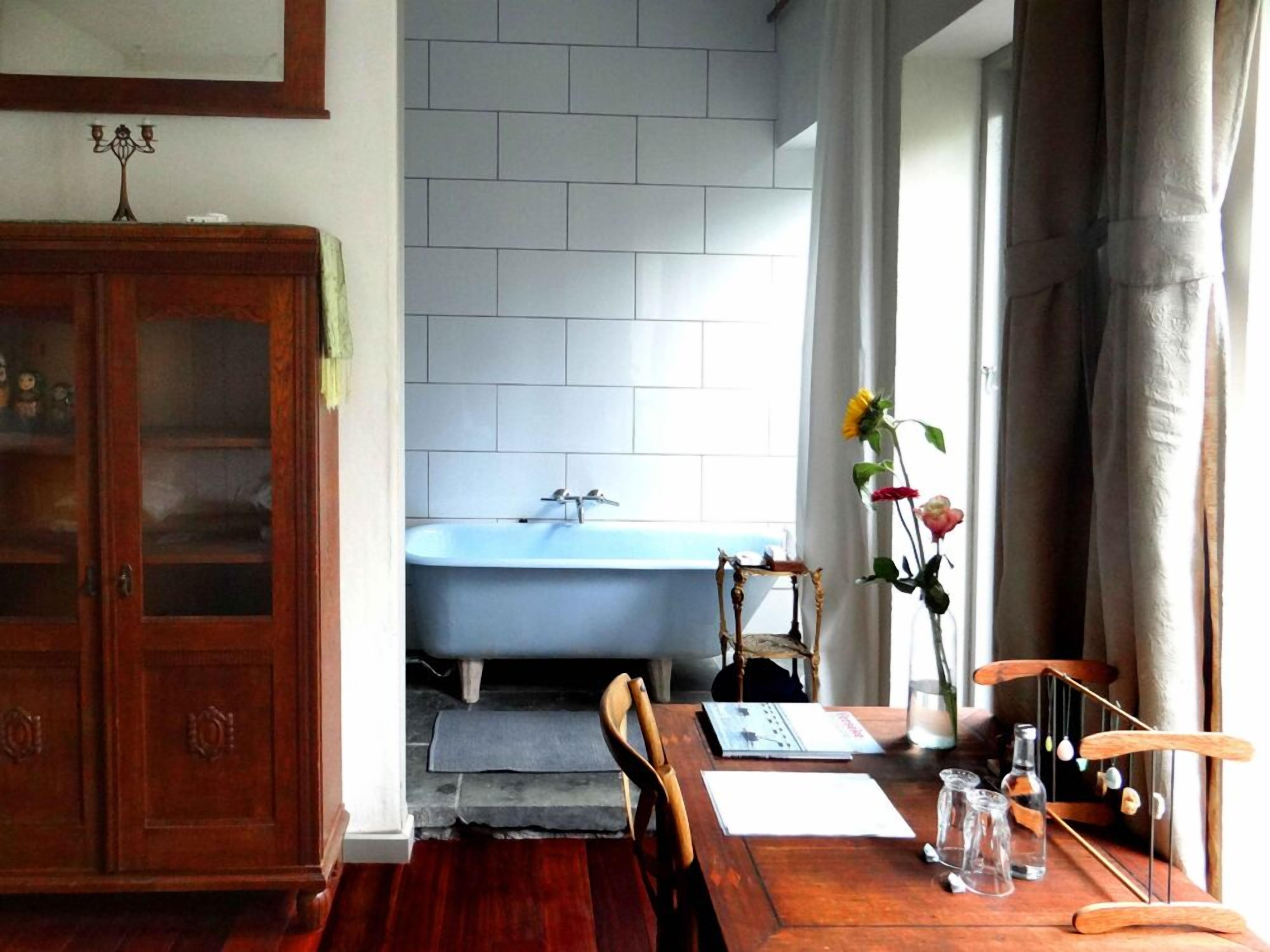 A room with a wooden cabinet, a blue bathtub, a wooden chair, a table with papers and bottles, and a window with curtains.