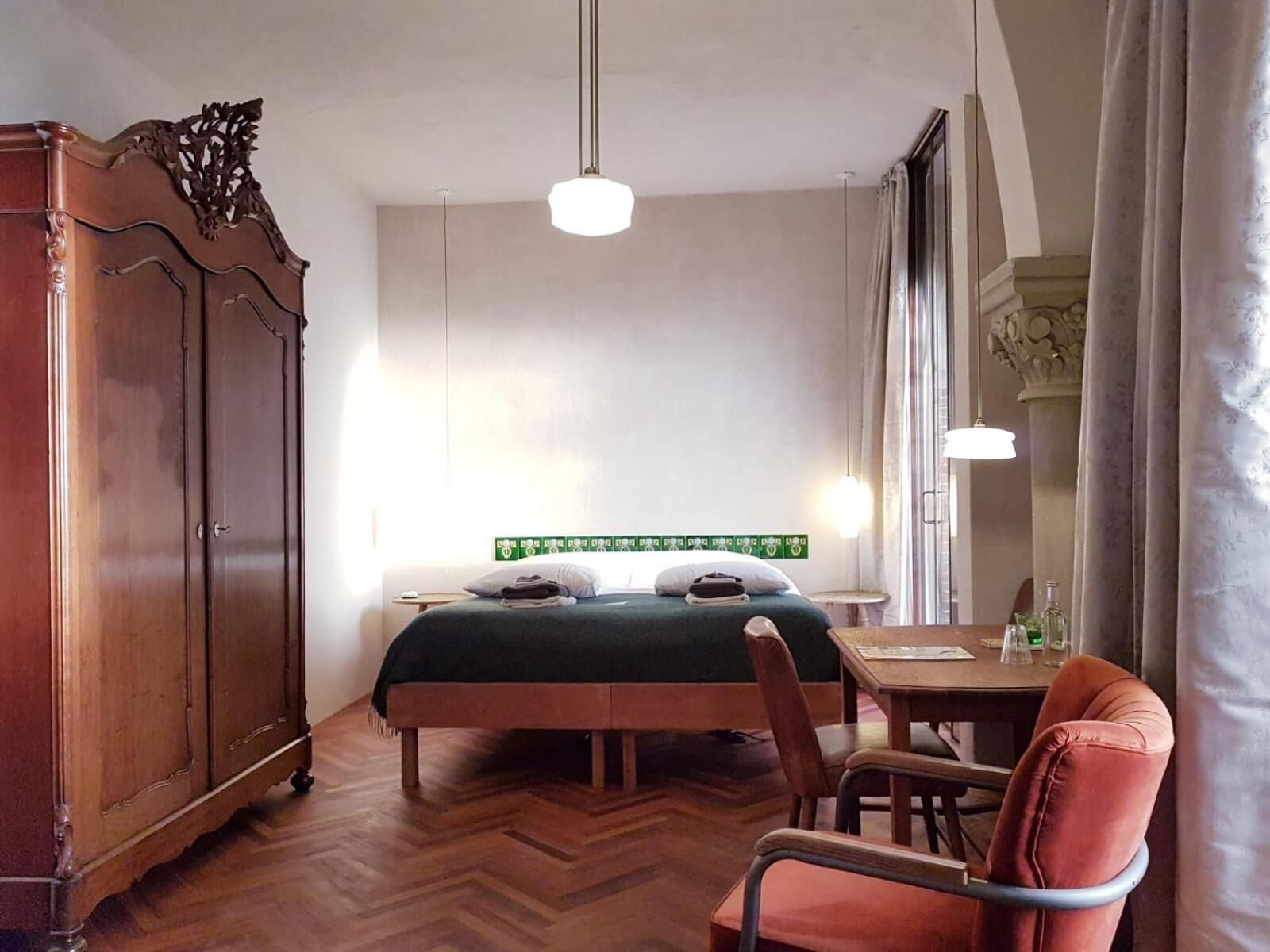 A bedroom with a bed, a wooden wardrobe, a desk with a chair, and a window with curtains.