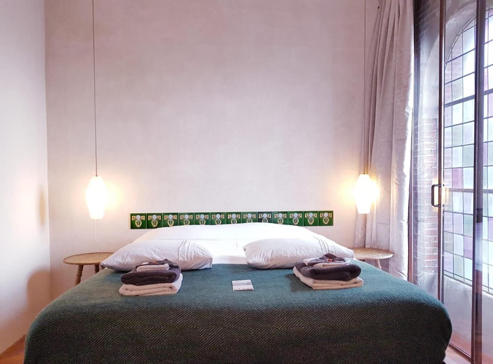 A hotel room with a bed, two bedside lamps, a window with curtains, and a wall with a green decorative panel.