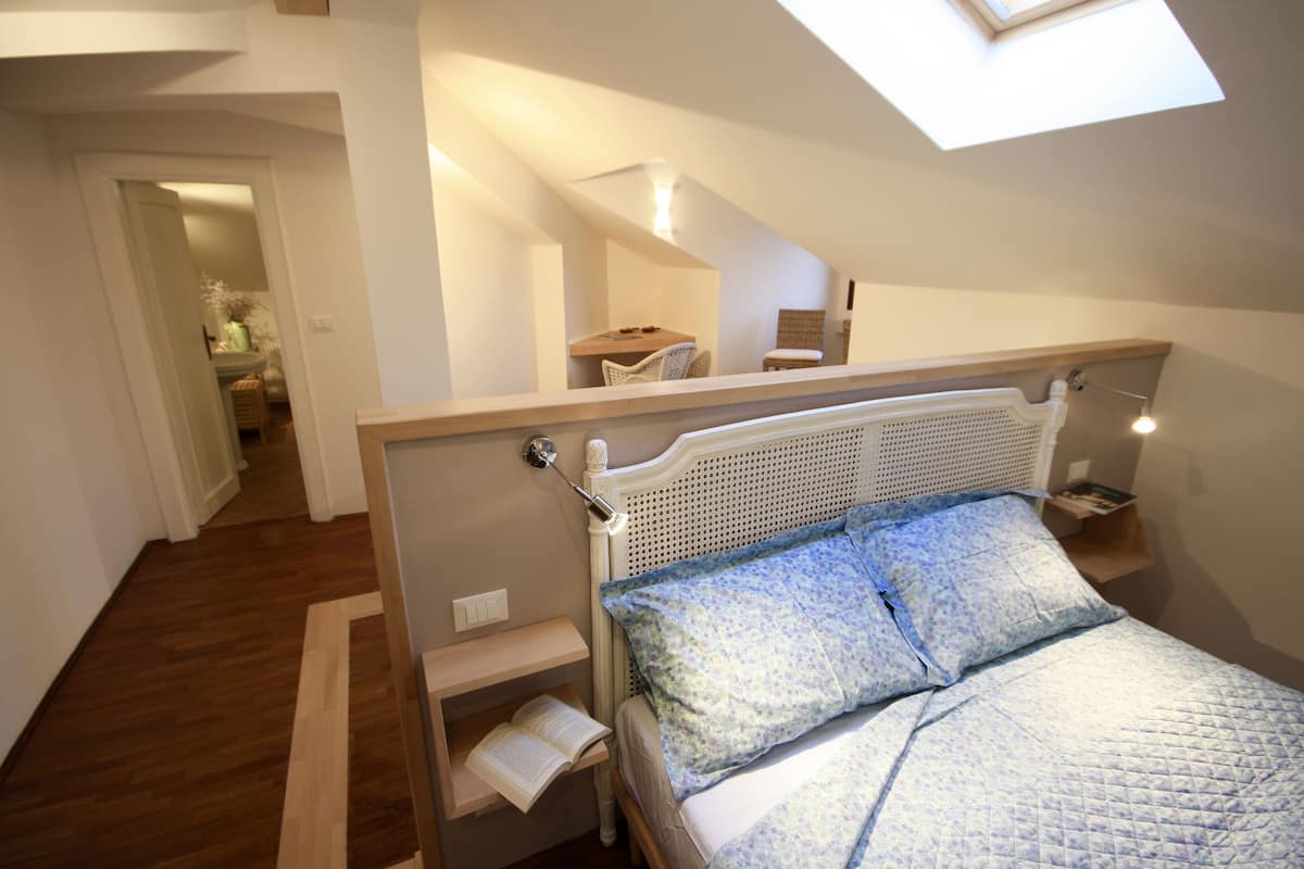 Une chambre avec une lucarne, un lit recouvert d’une courtepointe, une table de chevet avec un livre, et une petite table en bois avec une chaise à l’arrière-plan.