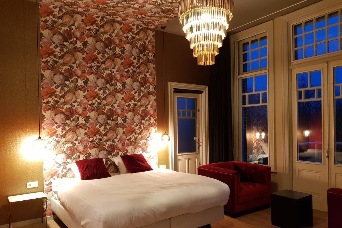 A bedroom with a large bed, a red sofa, a chandelier, and a floral wallpaper.