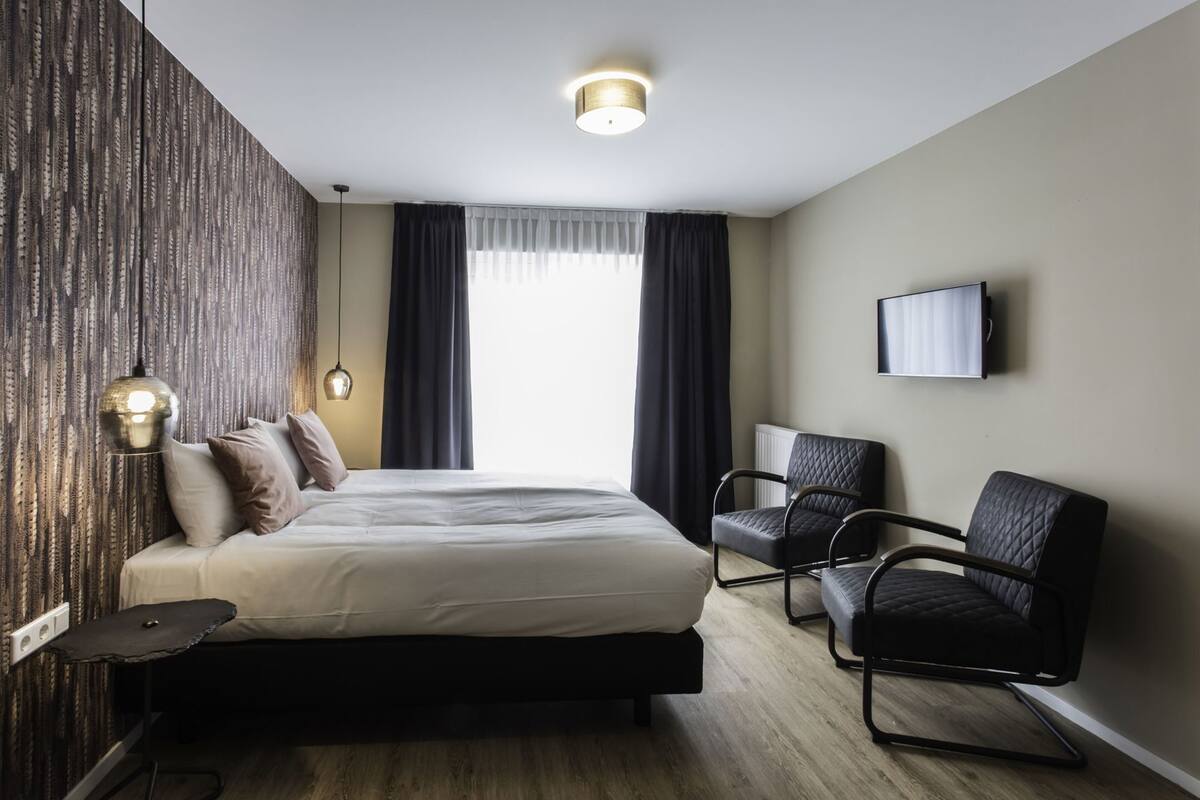 A hotel room with a bed, two chairs, a TV, and a window with curtains.