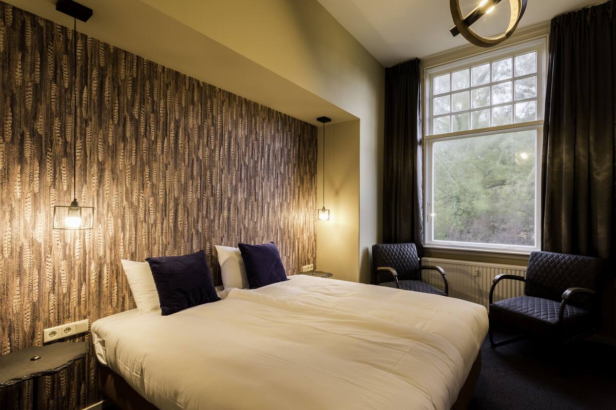 A hotel room with a large bed, two chairs, a window with curtains, and a patterned wall.