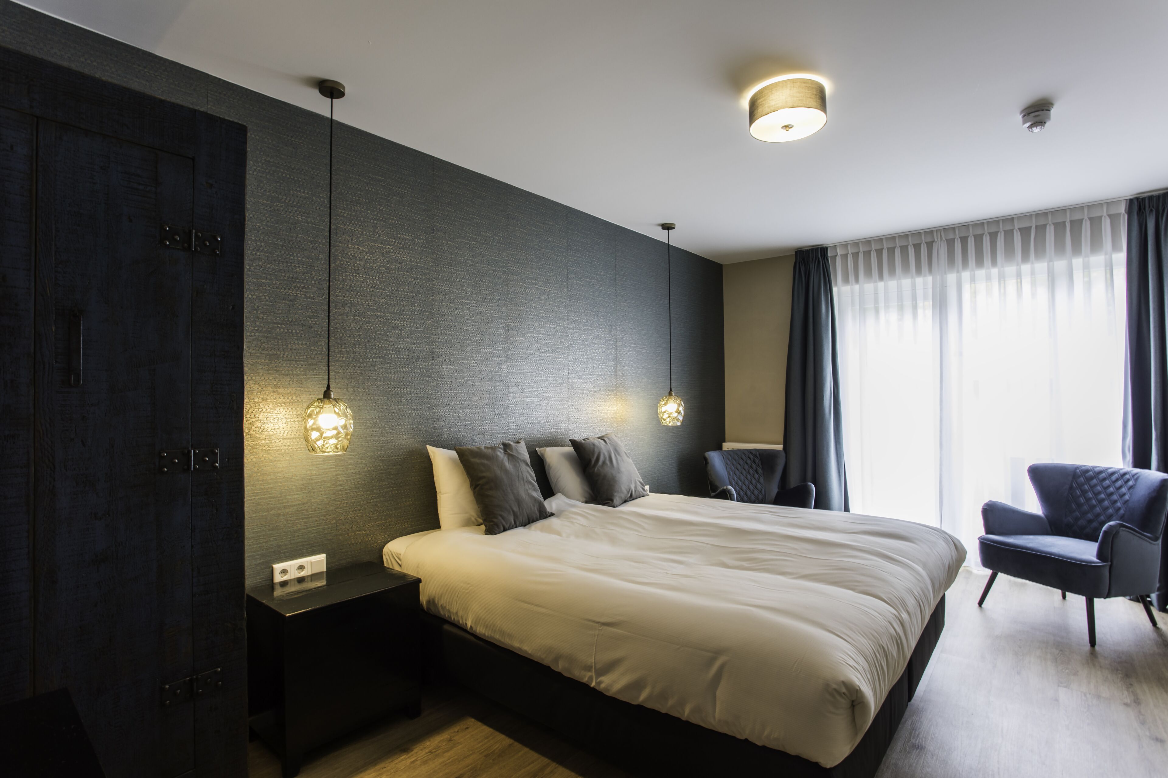 Una camera da letto moderna con un letto grande, pannelli in legno scuro, un comodino con una lampada e una sedia vicino alla finestra.