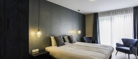 A modern bedroom with a large bed, dark wood paneling, a bedside table with a lamp, and a chair by the window.