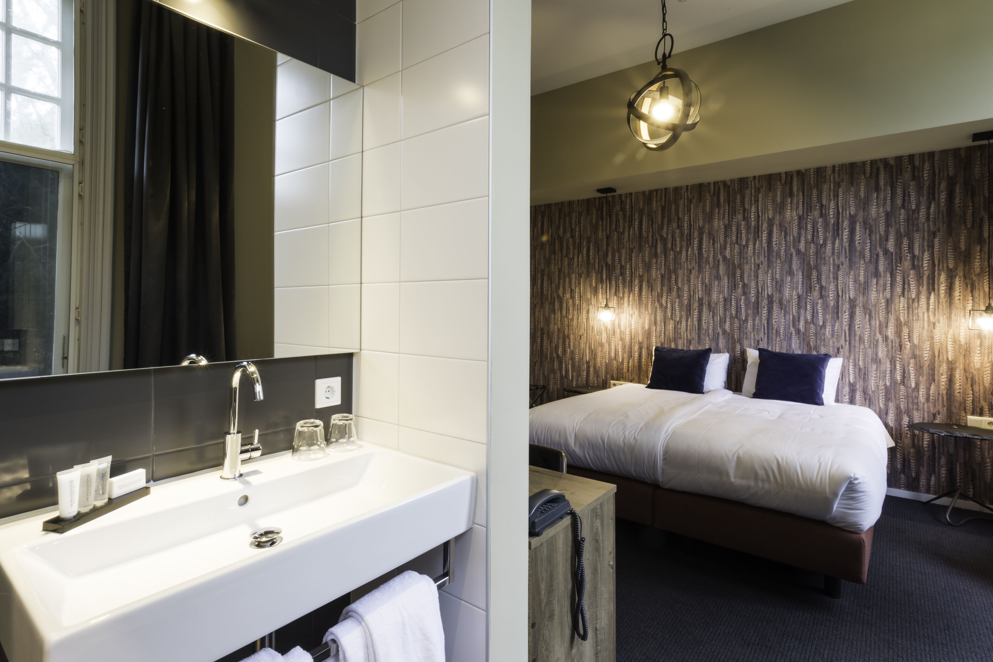Ein modernes Hotelzimmer mit einem großen Spiegel, einem weißen Waschbecken und einem ordentlich bezogenen Bett. Im Hintergrund ist eine Wand mit dunklem Muster zu sehen.