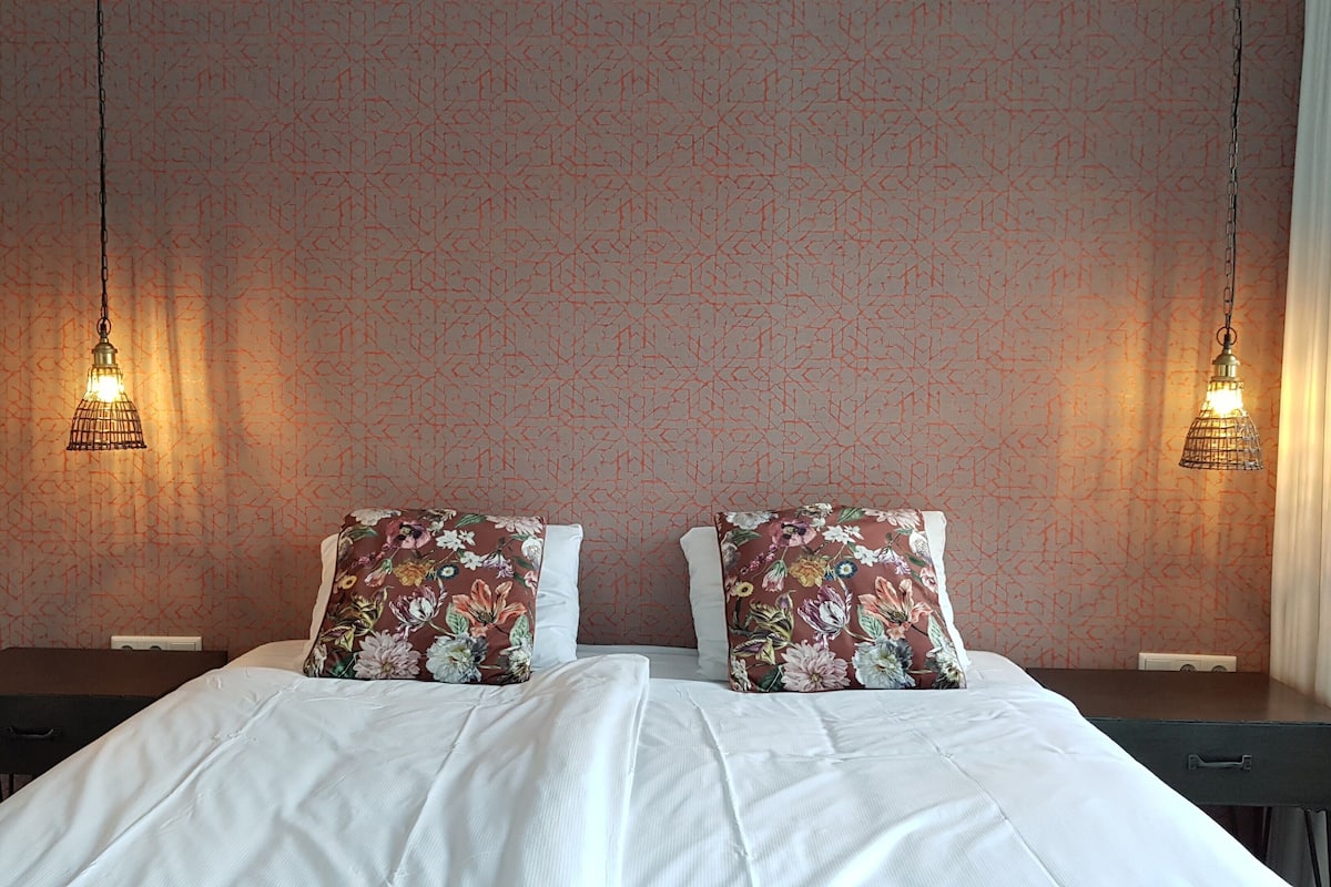 A neatly made bed with floral pillows, two bedside tables, and hanging light fixtures.