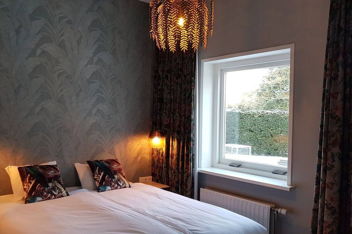 A bedroom with a bed, two bedside lamps, a window with curtains, and a wall with a leaf pattern.