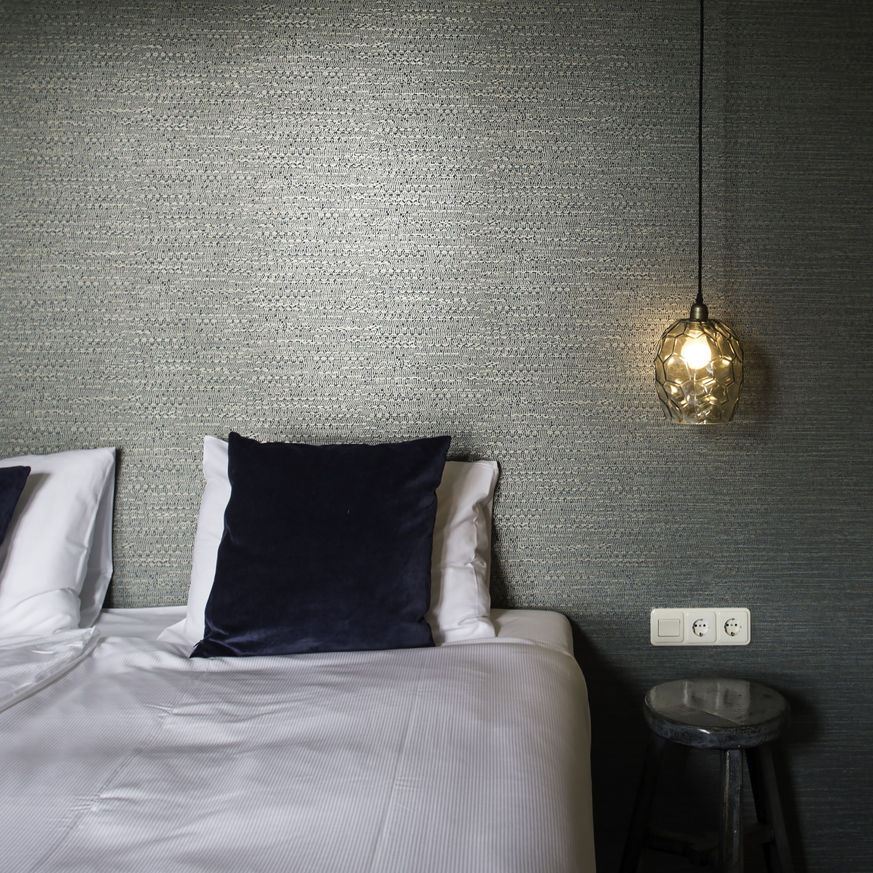 Een bed met witte en donkerblauwe kussens, een gestructureerde grijze muur, een hangende lamp en een klein rond tafeltje met een stopcontact.