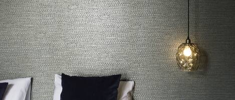 Un lit avec des oreillers blancs et bleu foncé, un mur gris texturé, un luminaire suspendu et une petite table ronde dotée d’une prise électrique.