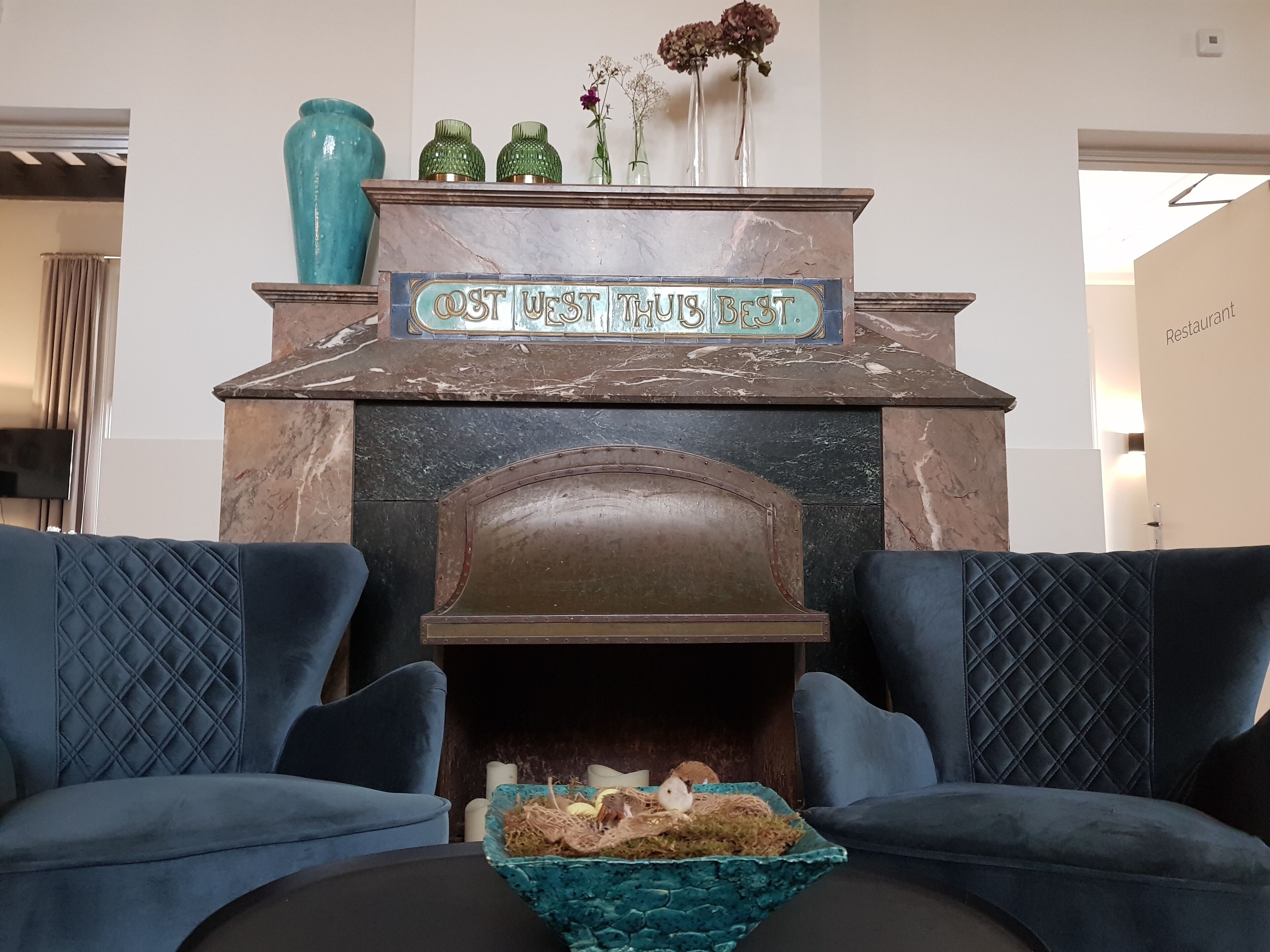 A fireplace with a sign reading "OST WEST TUS BEST" above it, flanked by two blue upholstered chairs.