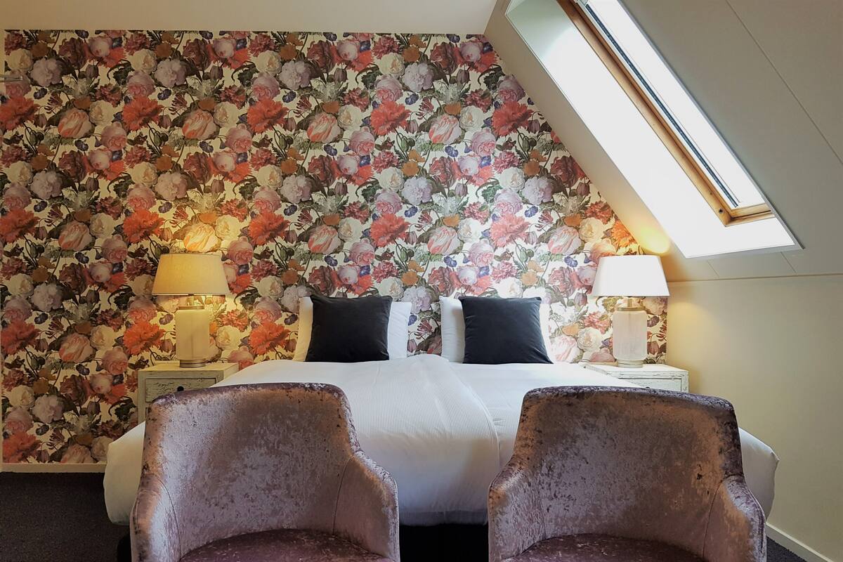 A hotel room with a floral wallpaper, a bed with white linens, two black pillows, a nightstand with a lamp, and two armchairs.