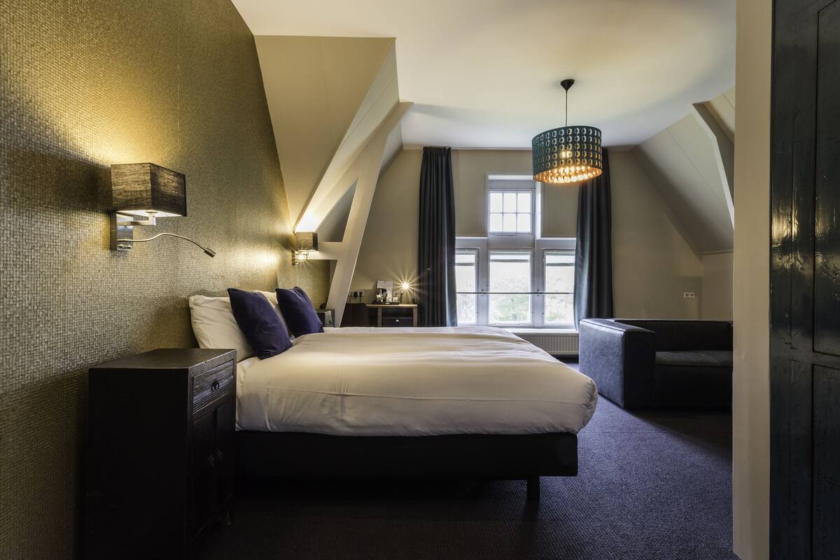 A modern hotel room with a large bed, a desk, a chair, a lamp, and a window with curtains.