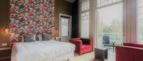 A modern bedroom with a large bed, a red armchair, a black chair, and a view of the outdoors through large windows.