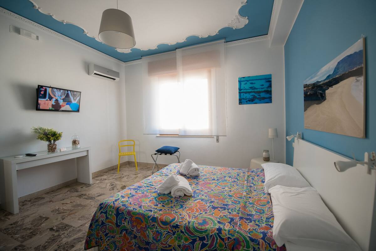 A bedroom with a bed, a desk, a chair, a TV, and artwork on the walls.