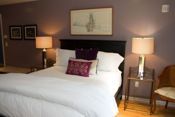A neatly made bed with white linens and a purple pillow, flanked by two bedside tables with lamps, a chair, and a framed artwork above the bed.
