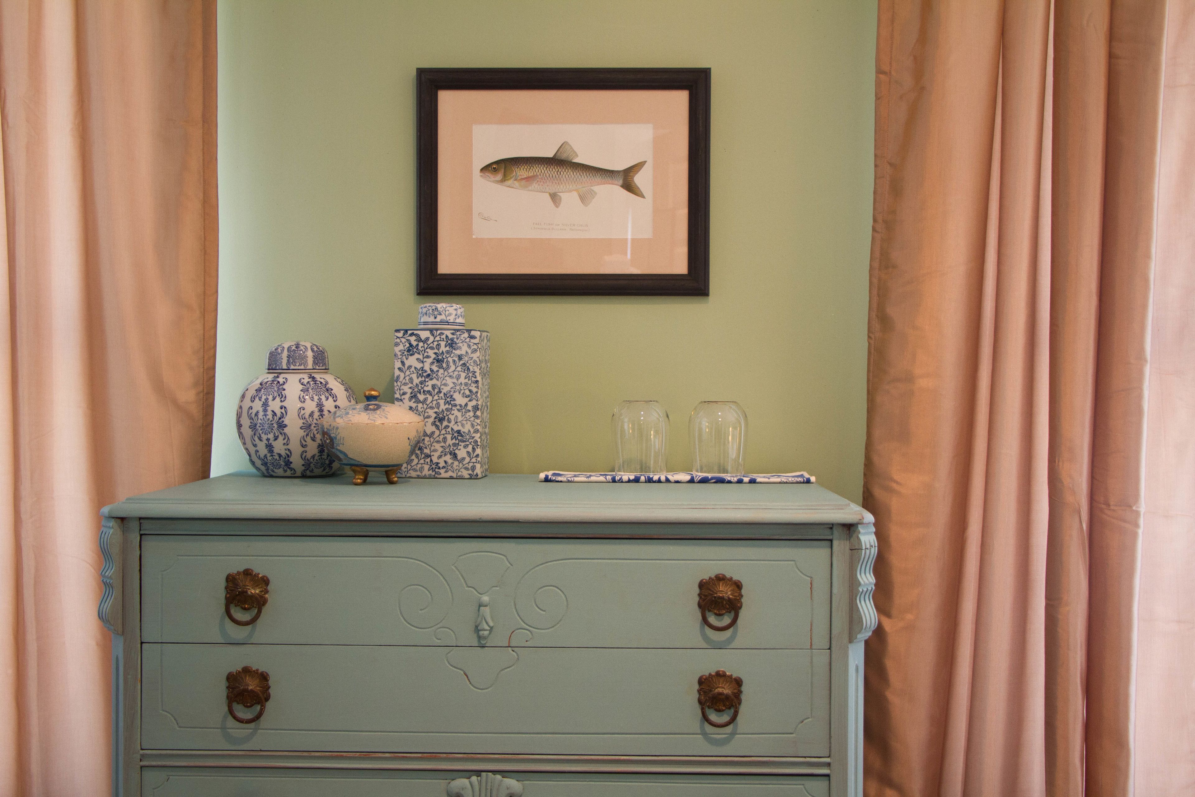 A light blue dresser with ornate handles, a framed fish illustration, and decorative vases.