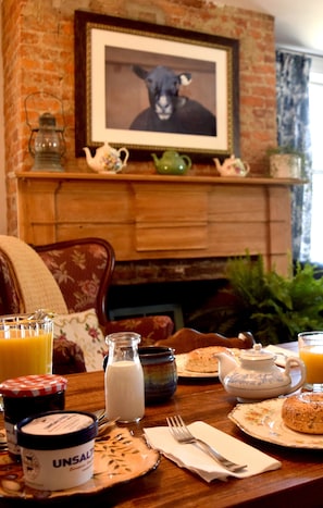 A breakfast table with a teapot, milk, and a bagel, set against a brick wall with a framed picture above.