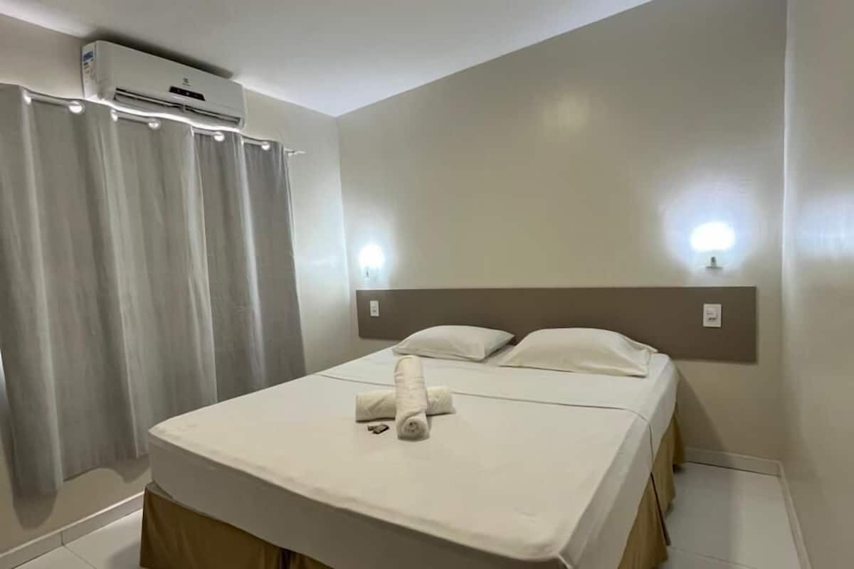 A hotel room with a bed, curtains, an air conditioning unit, and wall-mounted lights.