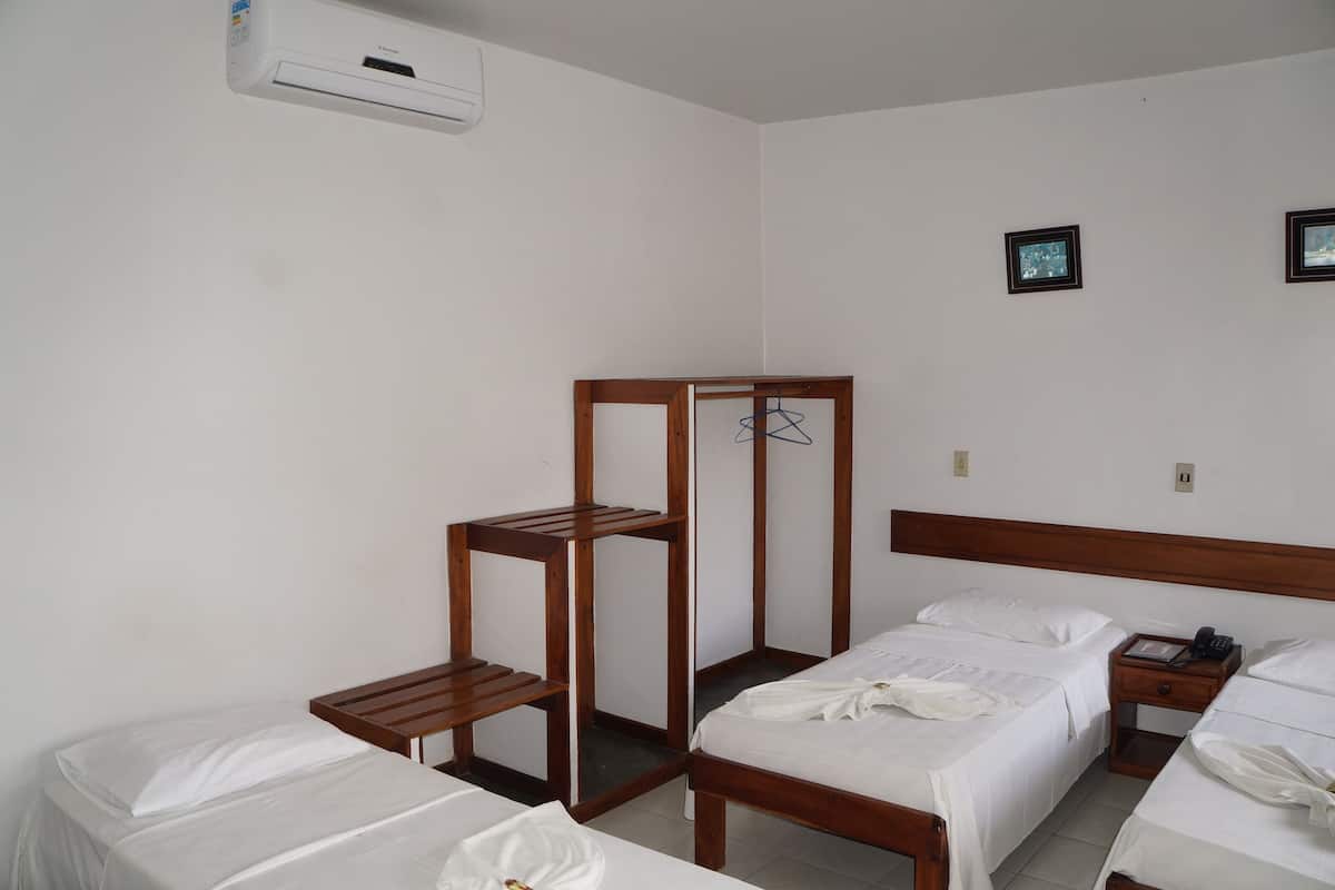 A hotel room with two beds, a wooden wardrobe, a bedside table with a lamp, and a wall-mounted air conditioner.