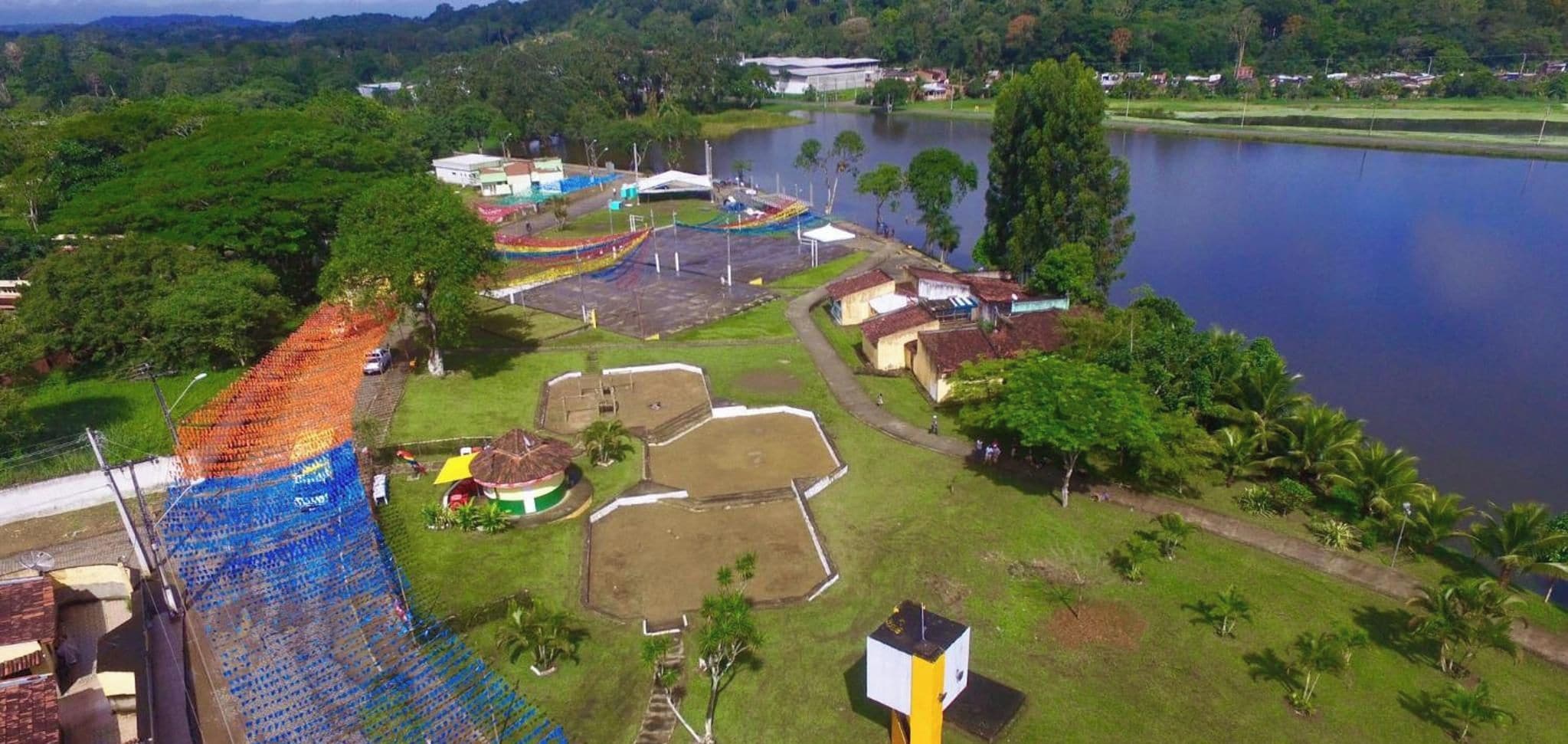 An aerial view of a property with a large body of water, a fenced area with colorful decorations, and several buildings with distinct architectural styles.