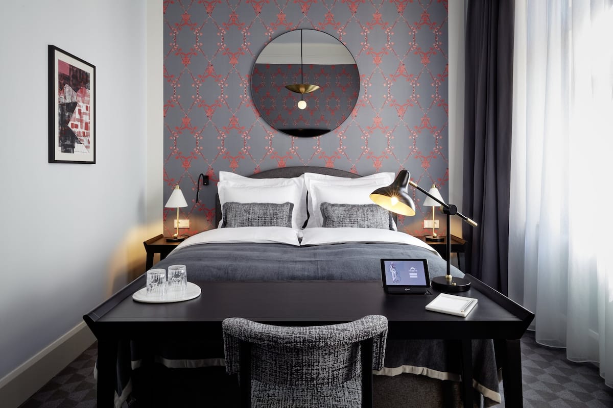 A modern hotel room with a large bed, a round mirror above it, two bedside tables with lamps, a desk with a chair, and a small TV on the desk.