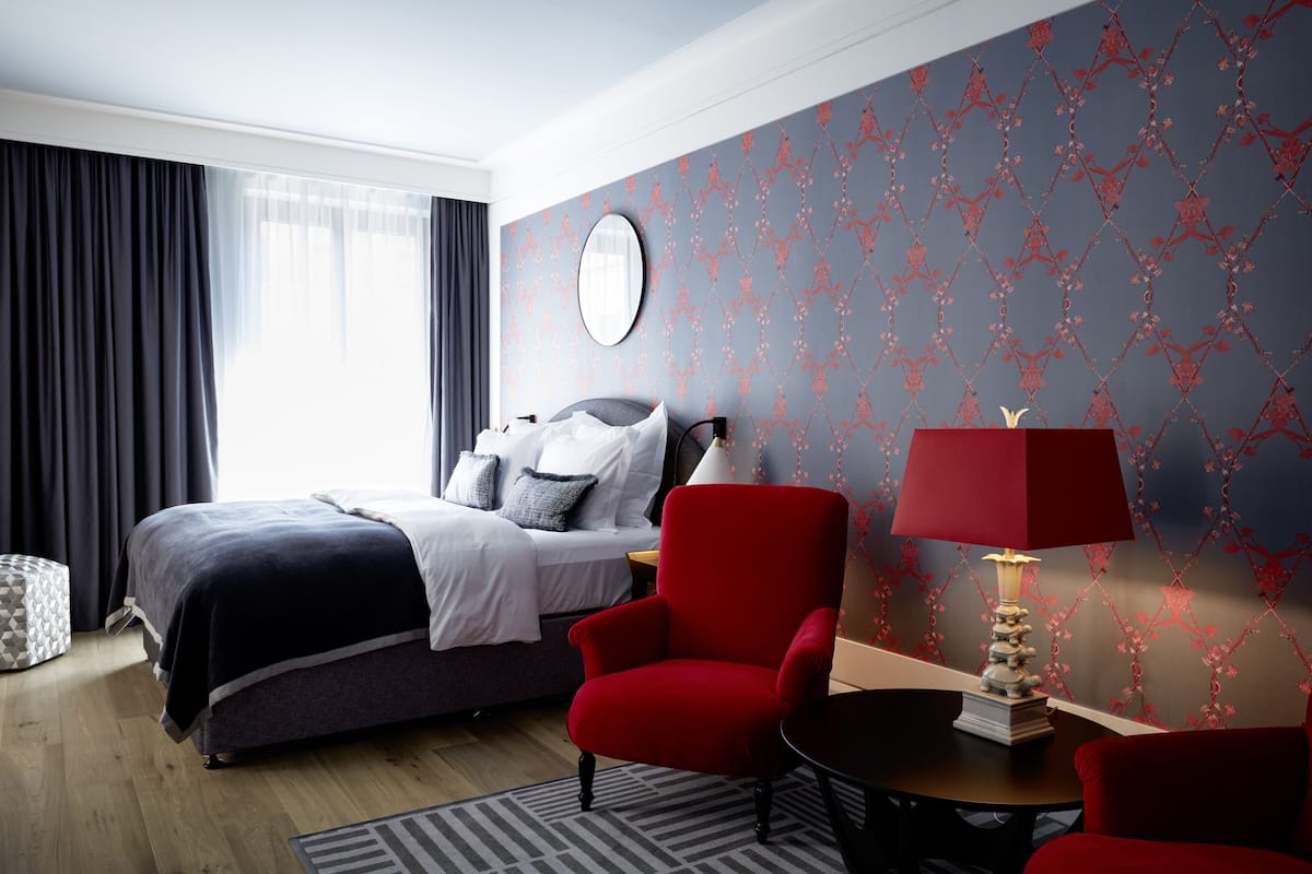 A modern hotel room with a bed, a red armchair, a round table with a lamp, and a patterned wallpaper.