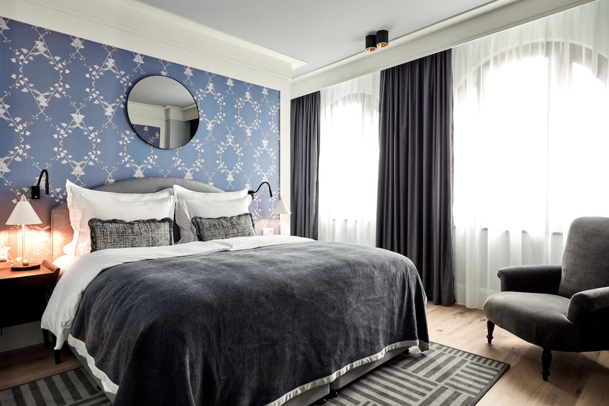 A bedroom with a large bed, a round mirror, a nightstand with a lamp, a chair, and a window with curtains.