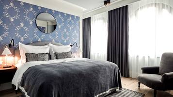 A bedroom with a large bed, a round mirror, a nightstand with a lamp, a chair, and a window with curtains.