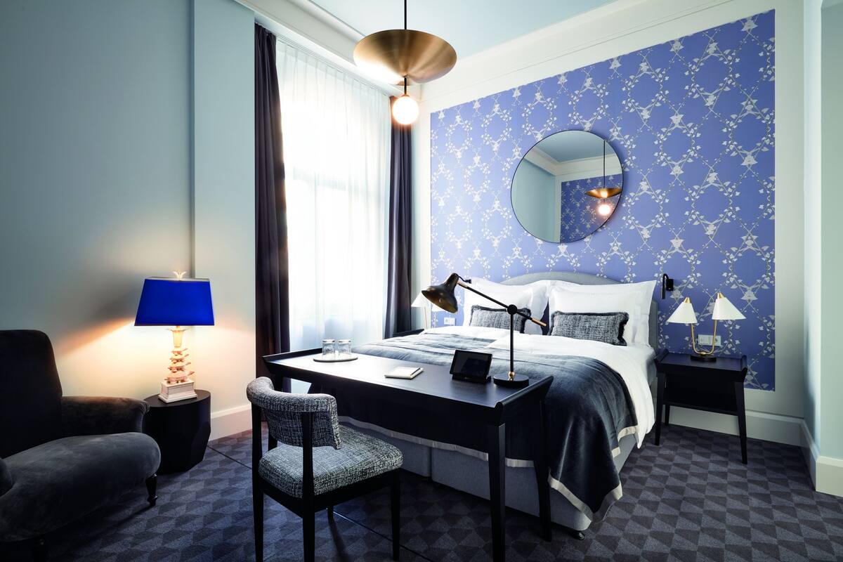 A hotel room with a bed, desk, chair, and a round mirror on the wall.