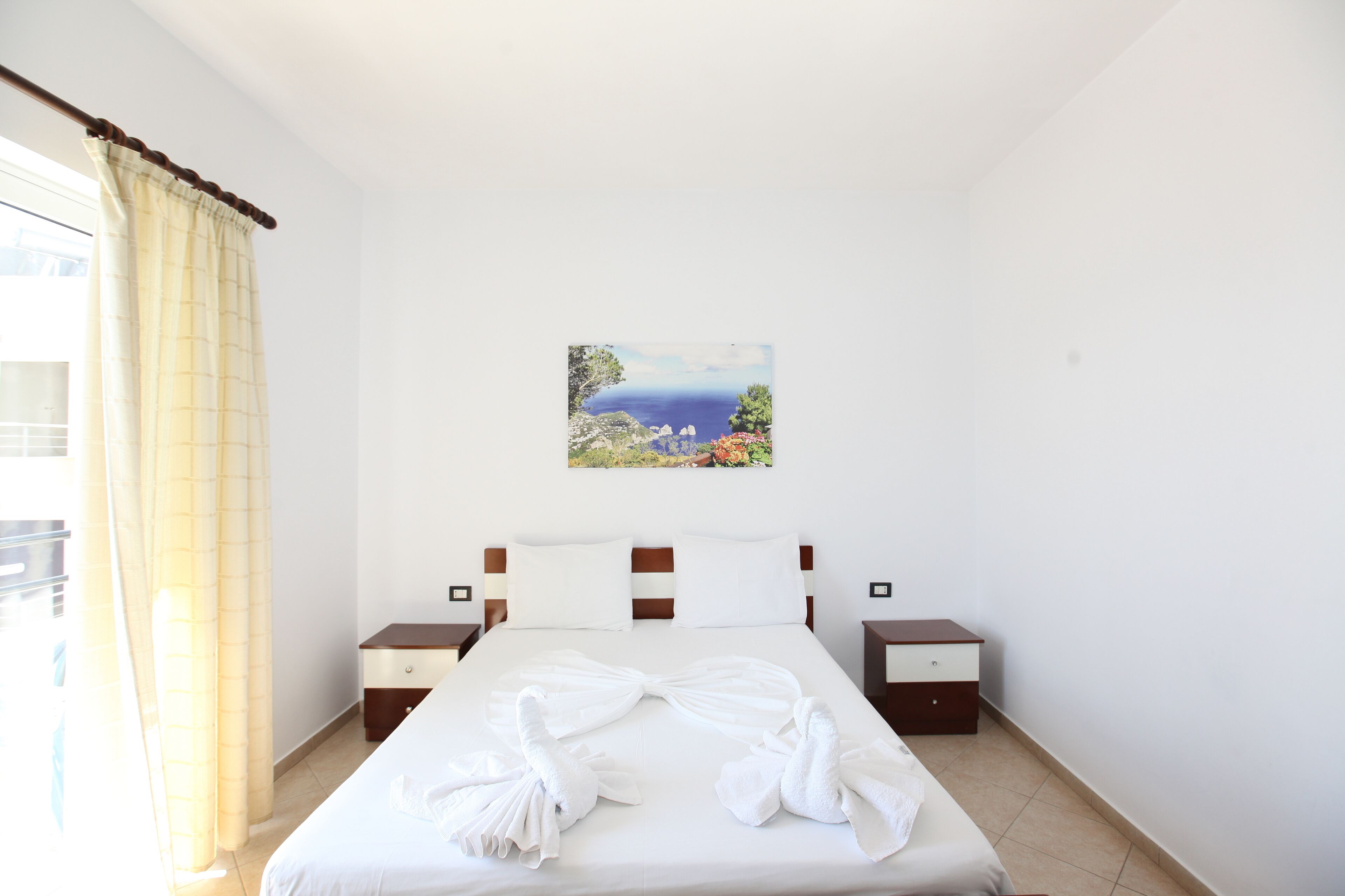A hotel room with a bed, a painting of a landscape, and a window with curtains.