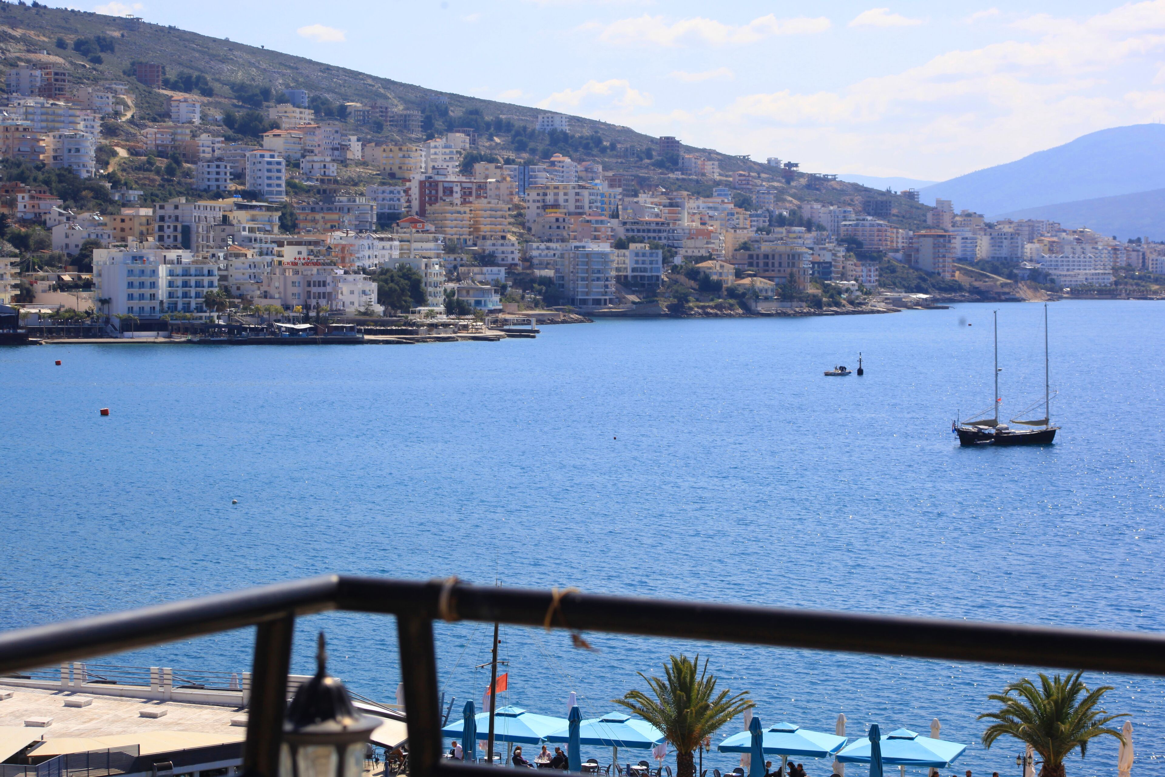 A coastal cityscape with buildings along the waterfront, a clear blue sea, and a sailboat.