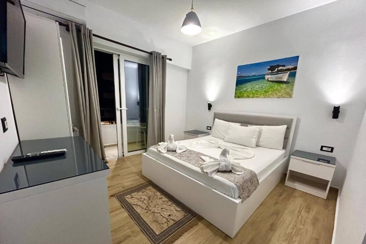 A modern hotel room with a large bed, a flat-screen TV, a bedside table, and a view of the sea through the window.