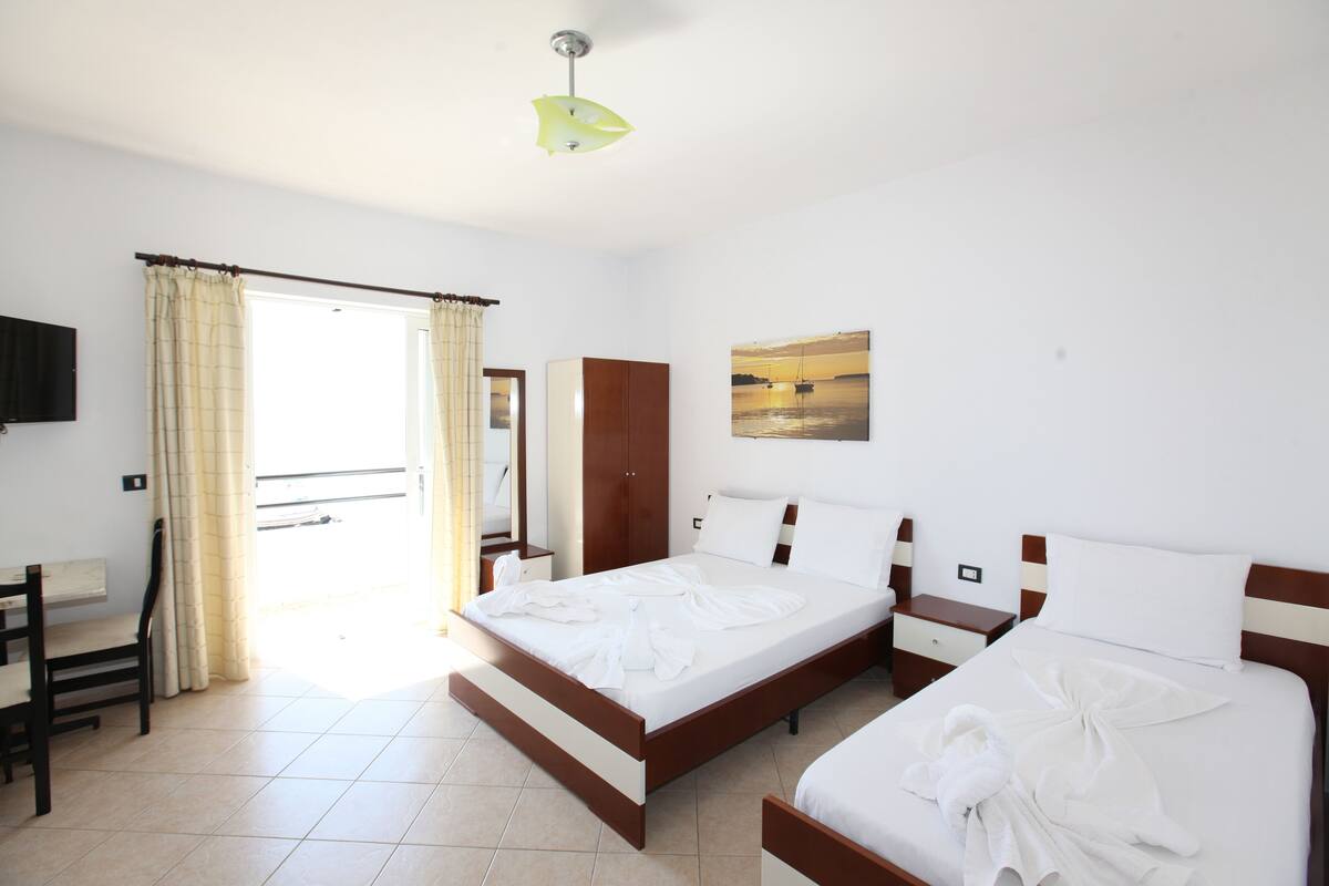 A hotel room with a large bed, a desk, and a chair. There is a window with curtains, a painting on the wall, and a ceiling light.
