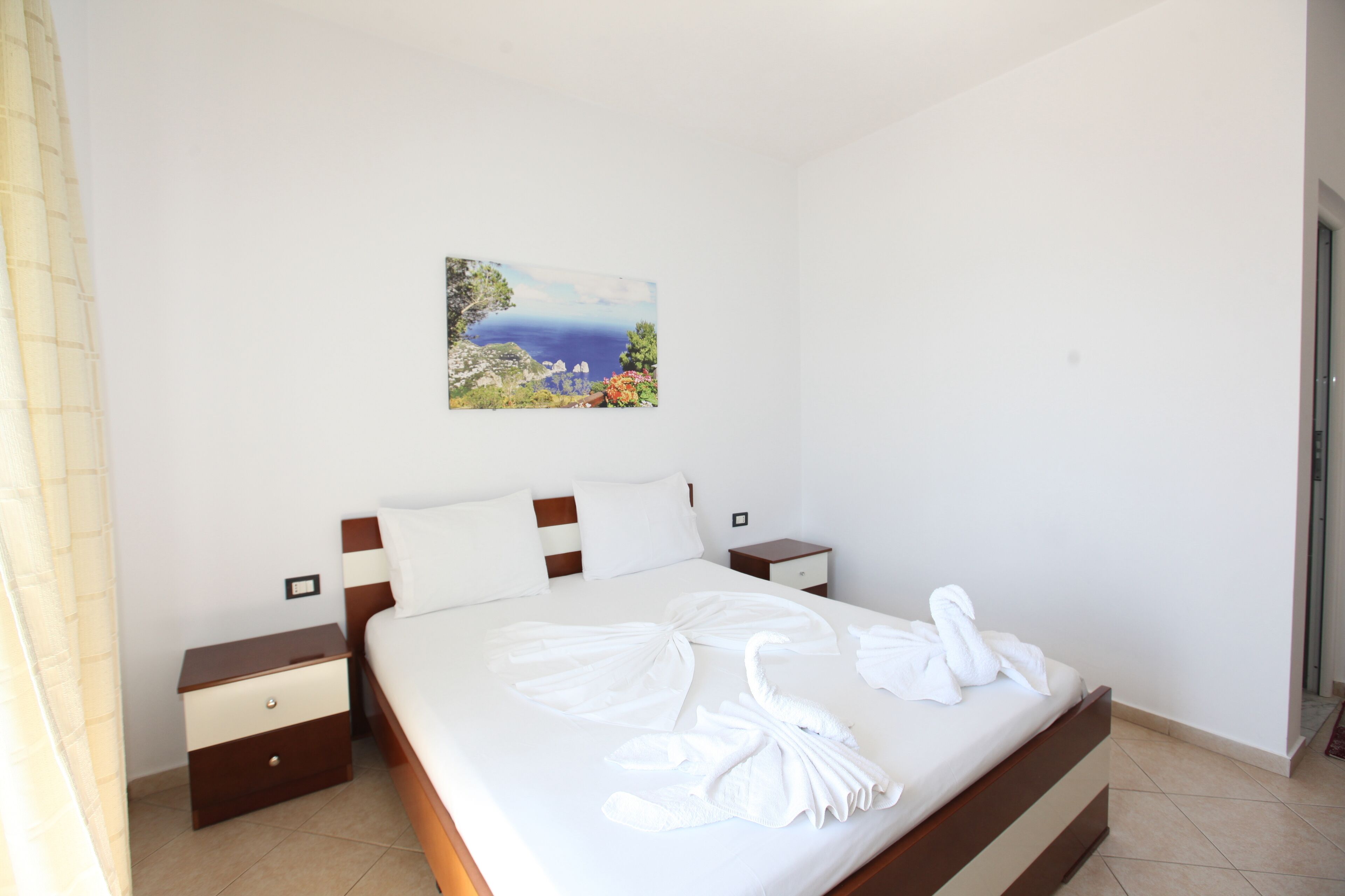 A hotel room with a bed, a nightstand, a drawer, a painting of a landscape, and a door.