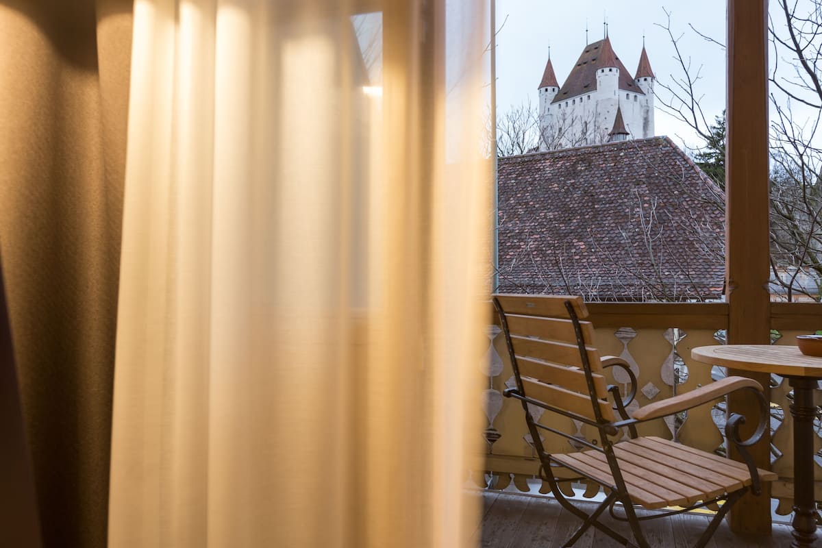 A balcony with a wooden chair and table, overlooking a castle with multiple towers.