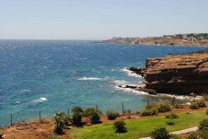 A coastal landscape with a rocky cliff, clear blue sea, and a distant town. - Andromachi Apartments (Rafina-Pikermi)