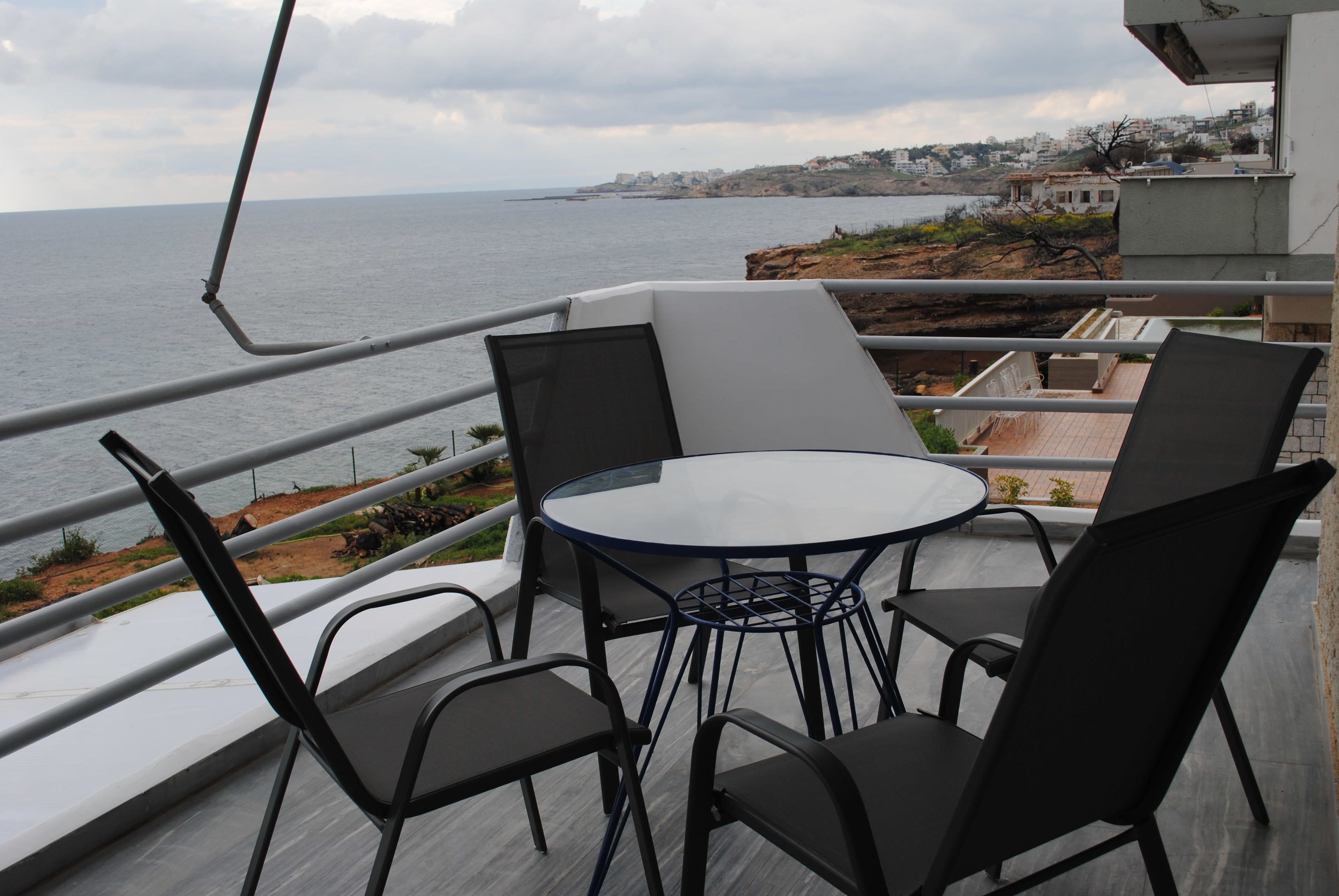A balcony with a round table and four chairs, overlooking a coastal area with buildings and cliffs.