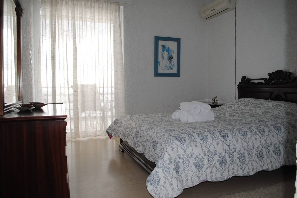 Une chambre à coucher avec un lit, une commode, un tableau encadré au mur et une fenêtre avec des rideaux légers.
